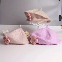 Trois bérets fille en maille beige, rose pâle et lilas, ornés de fleurs roses, présentés ensemble.