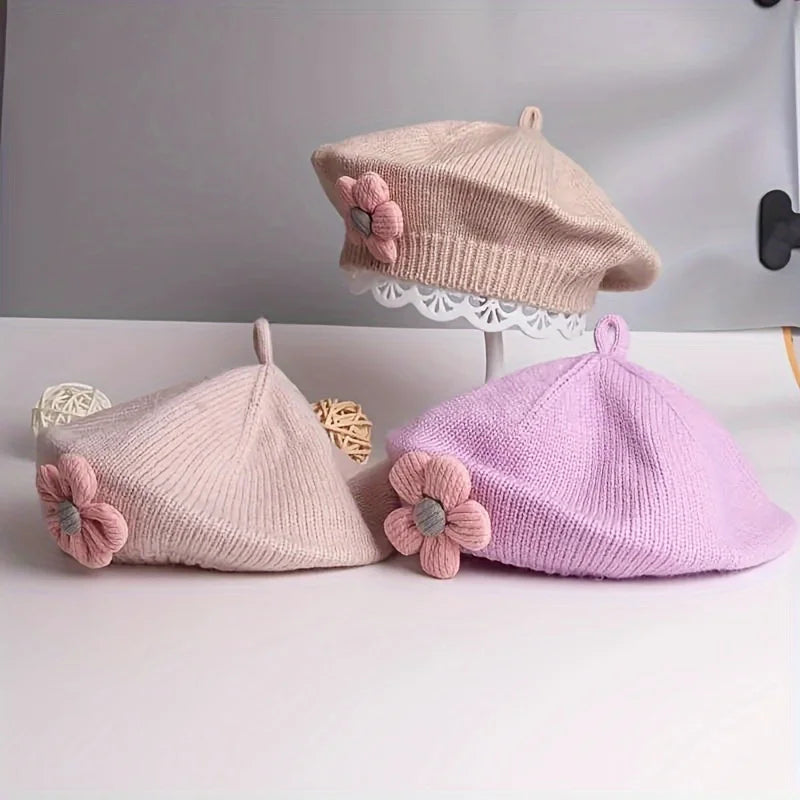 Trois bérets fille en maille beige, rose pâle et lilas, ornés de fleurs roses, présentés ensemble.
