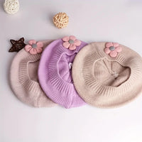Trois bérets fille en maille rose pâle, lilas et beige, vus de l’intérieur avec fleurs décoratives.